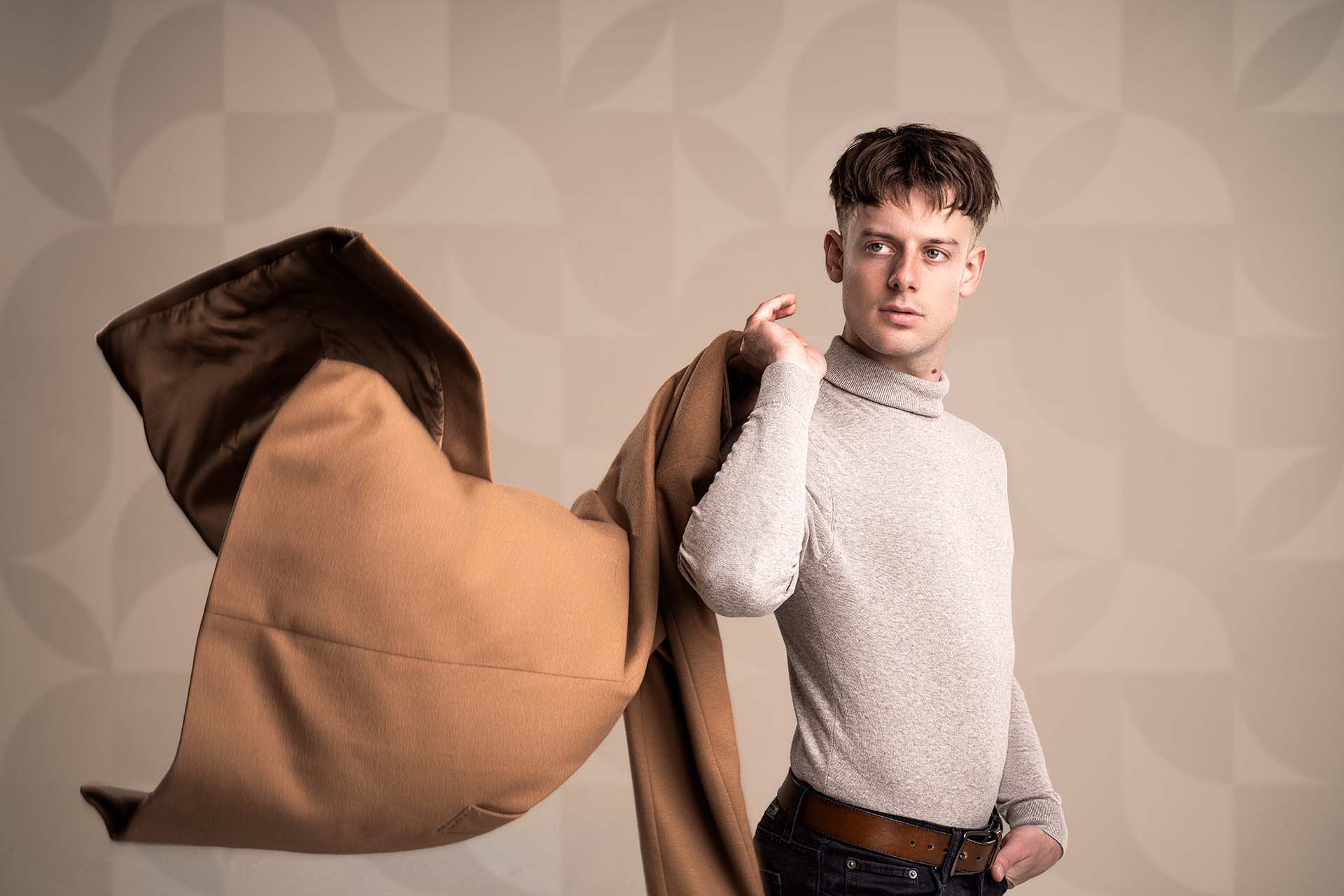Male model walking dynamically in a tan coat against a patterned backdrop