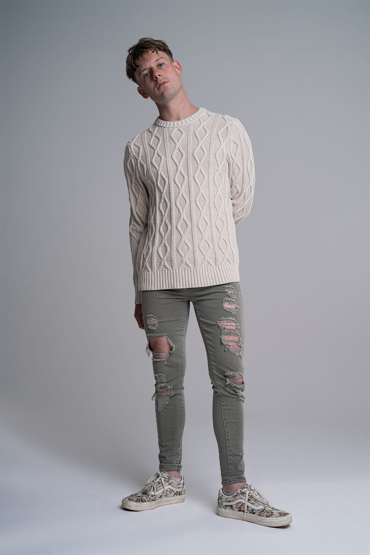 Alt: Full-body portrait of a male model in a cream cable-knit sweater and distressed jeans, studio photography