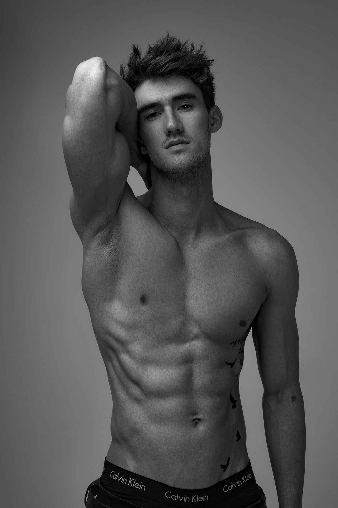 Black-and-white photo of a shirtless male model facing forward with his arm raised behind his head, studio portrait