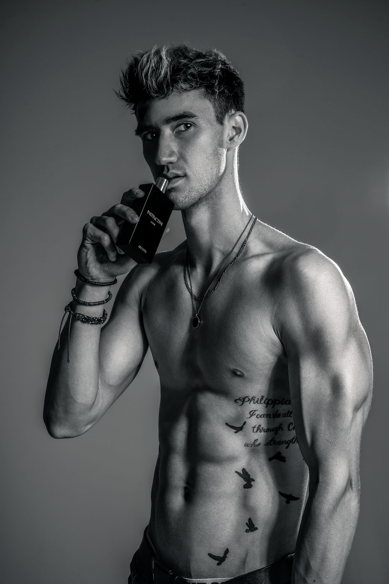 Black-and-white shirtless portrait of a male model holding a fragrance bottle near his face