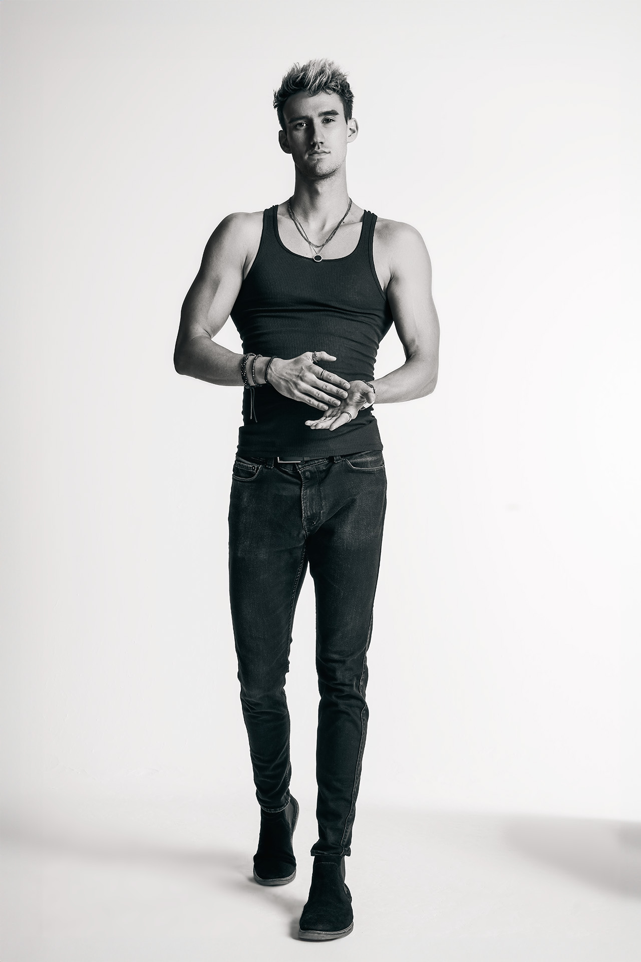 Full-body portrait of a male model in a black tank top and dark jeans, hands clasped at waist, wearing a watch