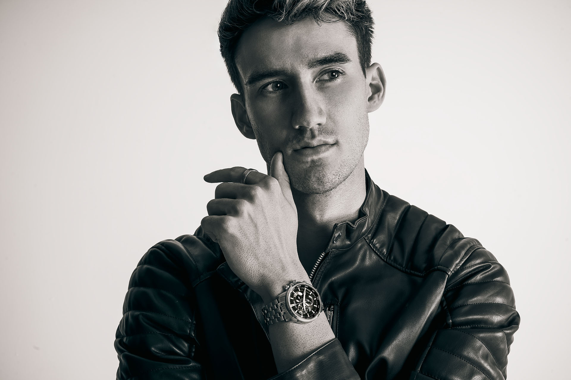 Black-and-white close-up portrait of a male model in a leather jacket, hand resting near his chin, wearing a watch