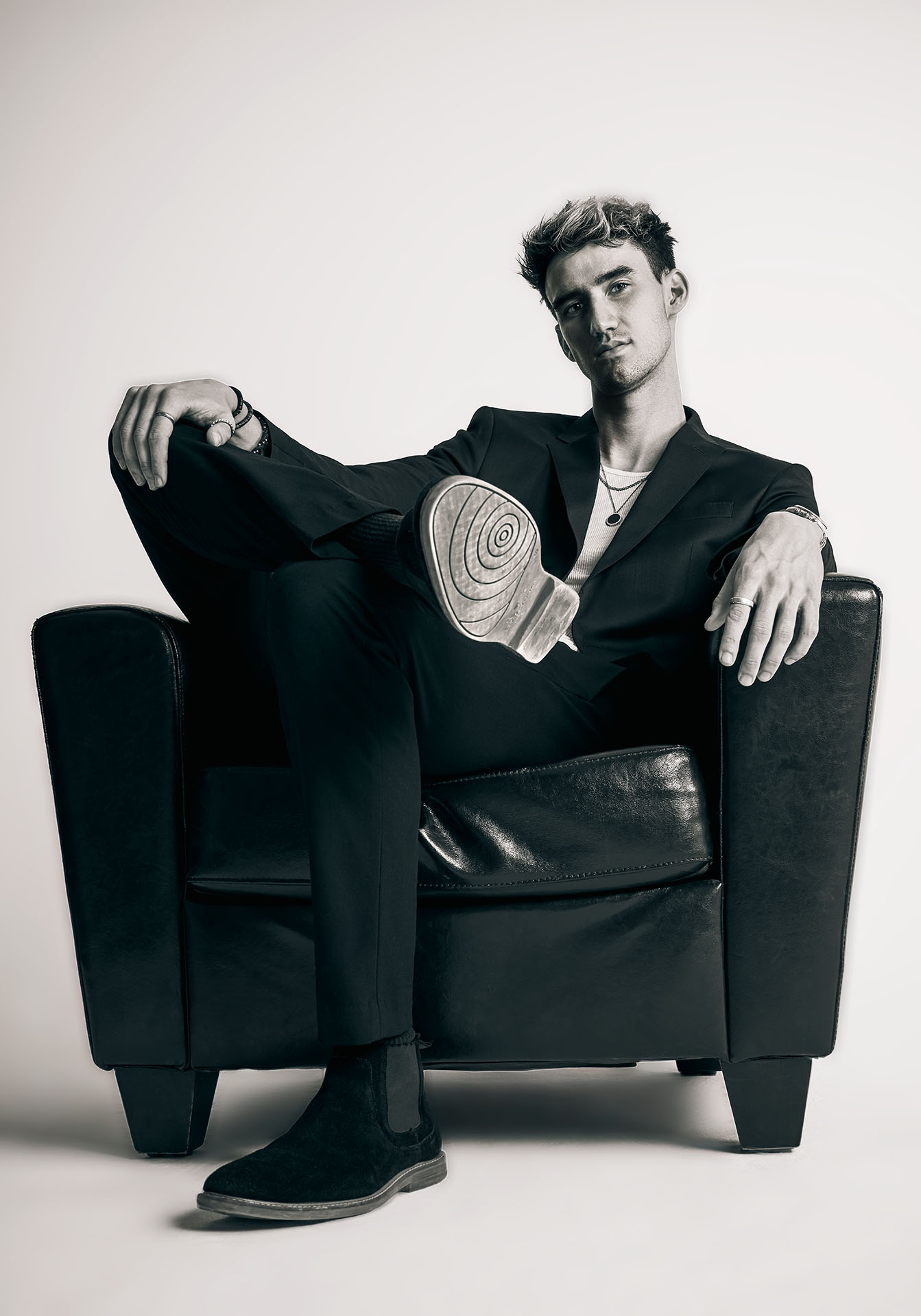 Black-and-white portrait of a male model seated on a chair with hand on knee and side of chair