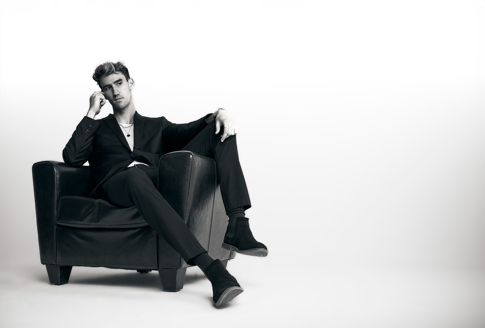 model in a suit seated in a leather armchair with legs draped over the armrest, hand raised near his eye