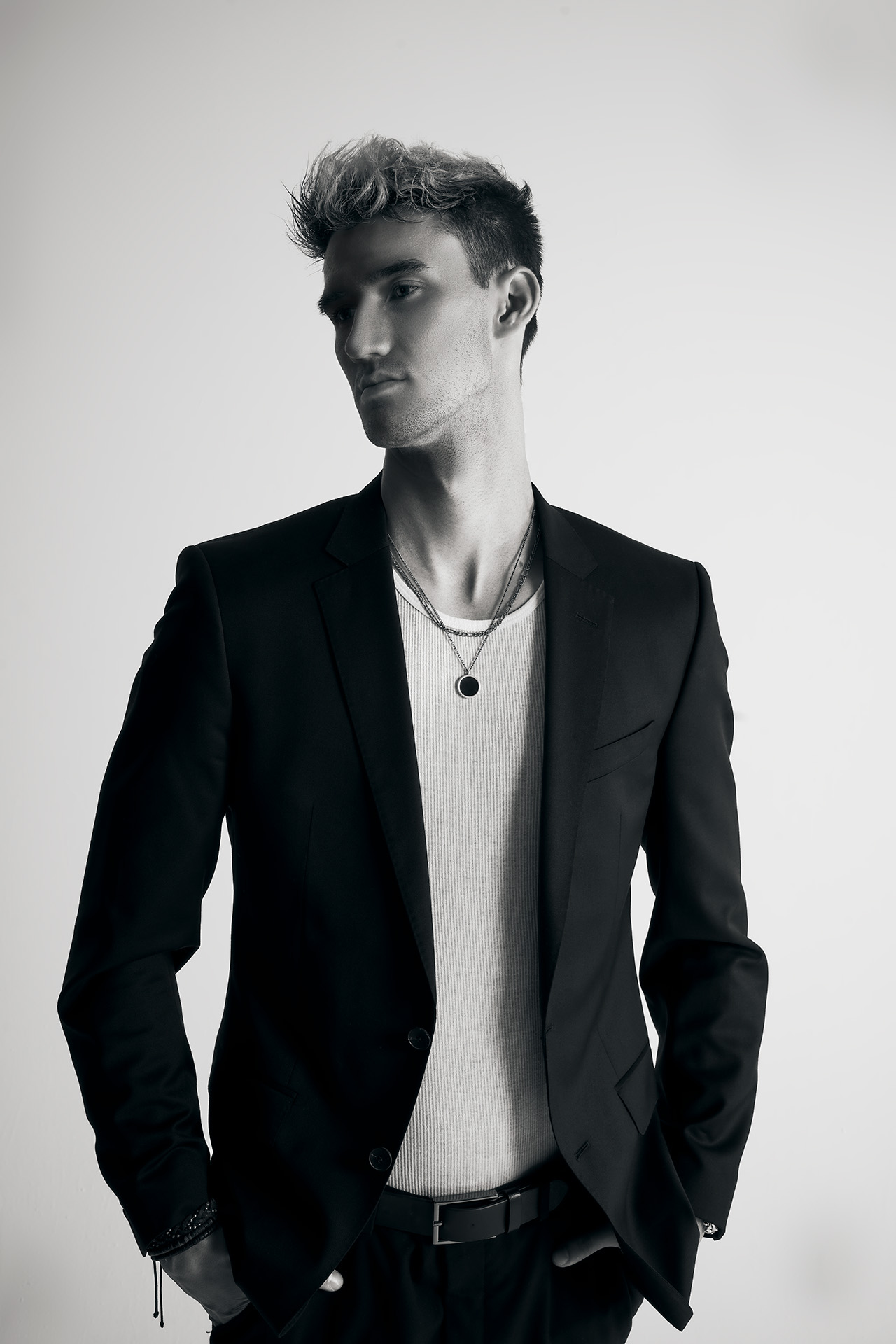 Black-and-white standing portrait of a male model in a black blazer with hand in pocket, wearing a necklace