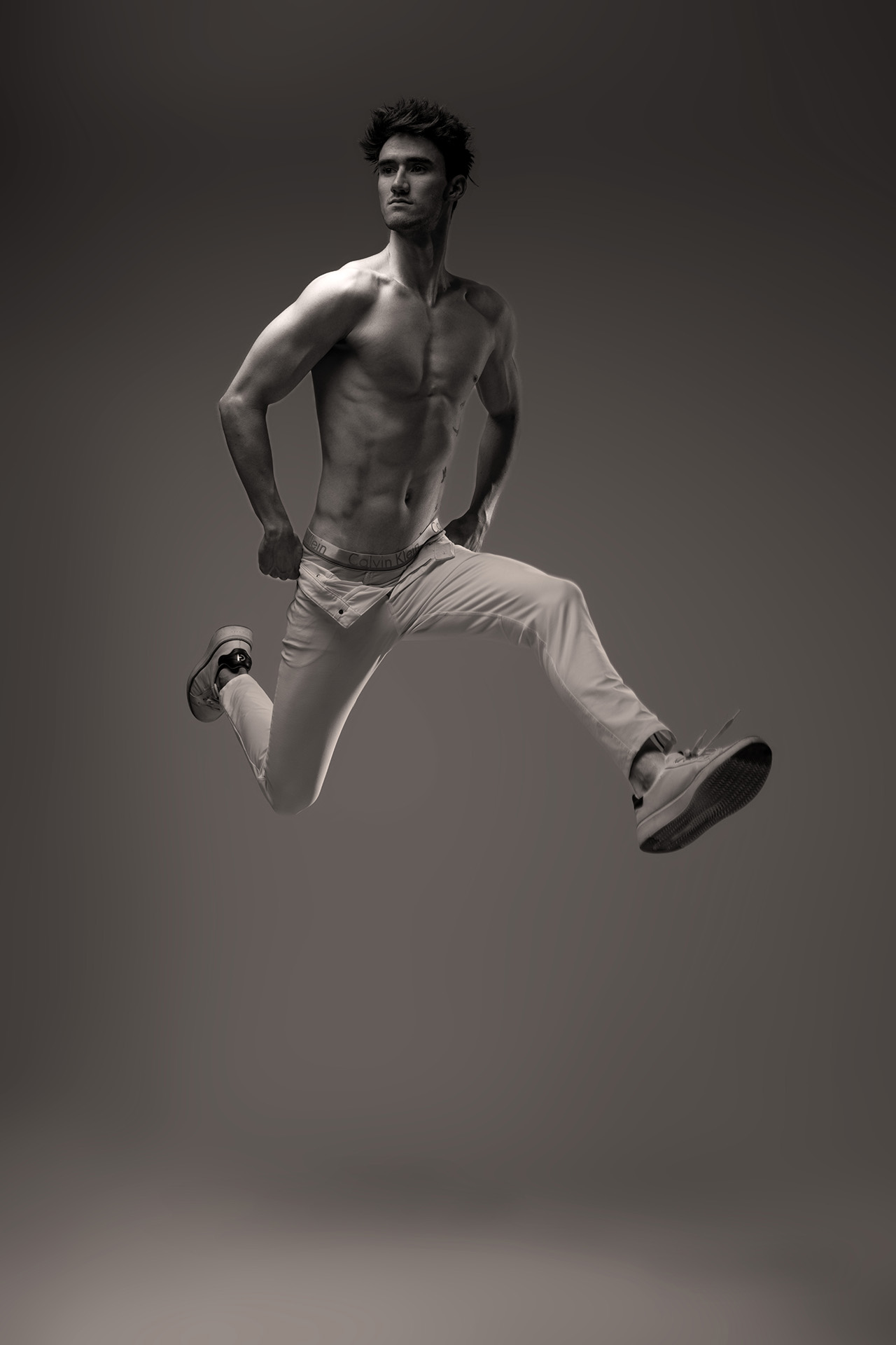 Shirtless male model leaping in white pants, studio portrait
