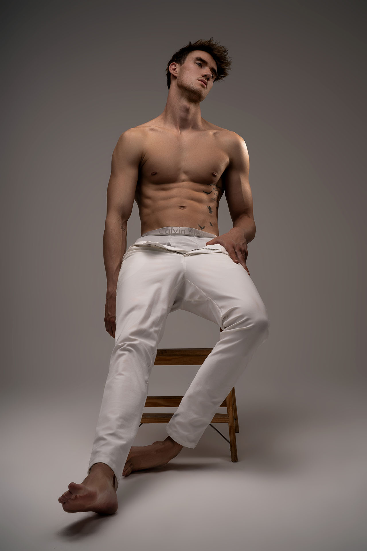 Shirtless male model seated on a stool with one leg raised, gray studio background