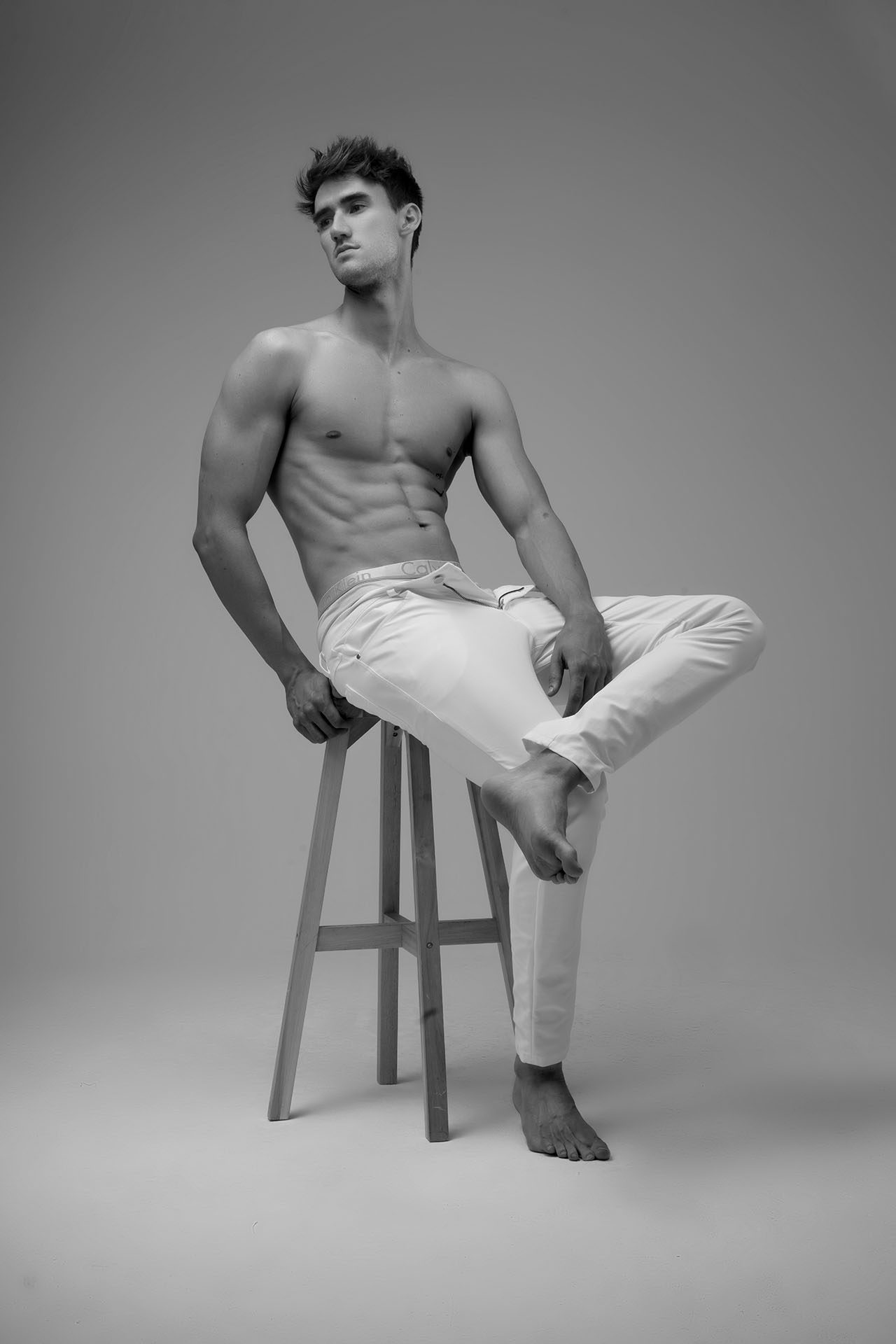 Shirtless male model seated on a stool with one foot propped up, white pants, studio portrait