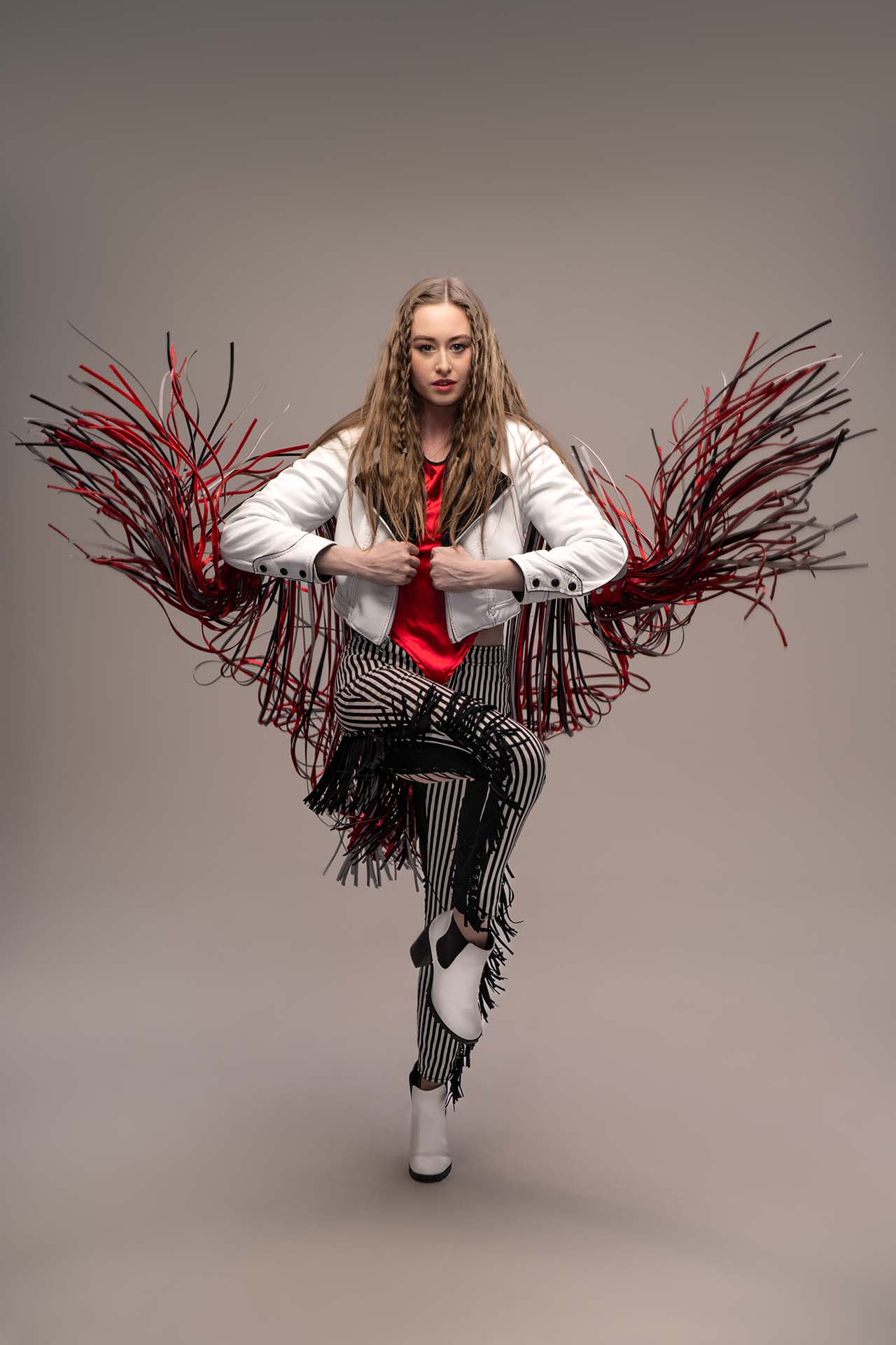 Female model in an elaborate red and black and red fringe coat over striped pants, dynamic crouched pose