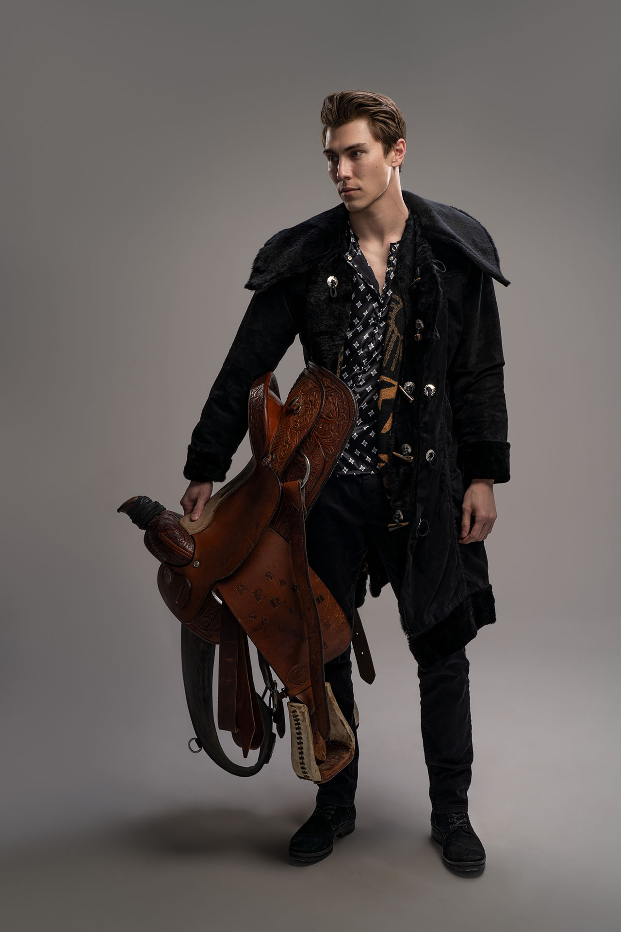 Man in a dark patterned coat holding a saddle prop, western-themed editorial photography