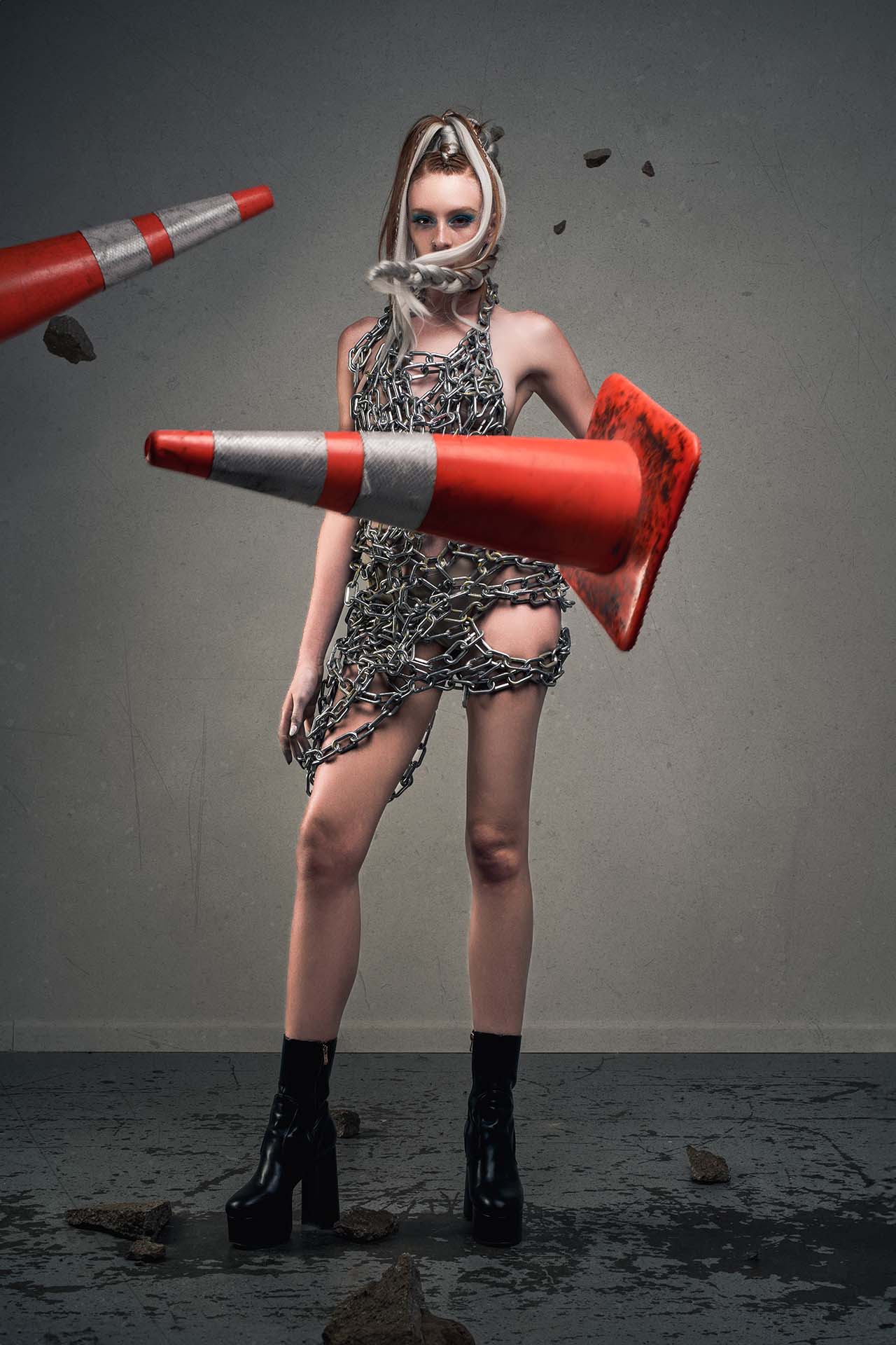 Model in a chain-link dress with a traffic cone flying through the air and a second cone flying overhead, dramatic avant-garde editorial photography