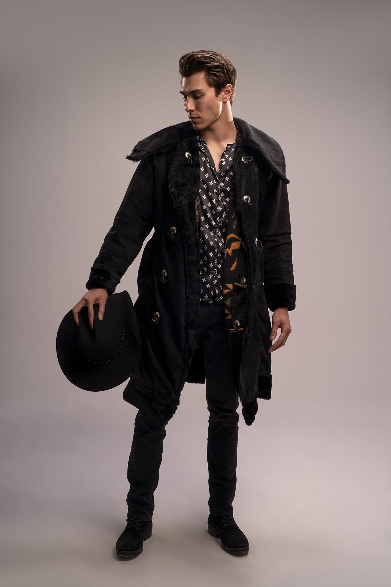 Man in a dark patterned coat holding a black hat, western-themed editorial portrait