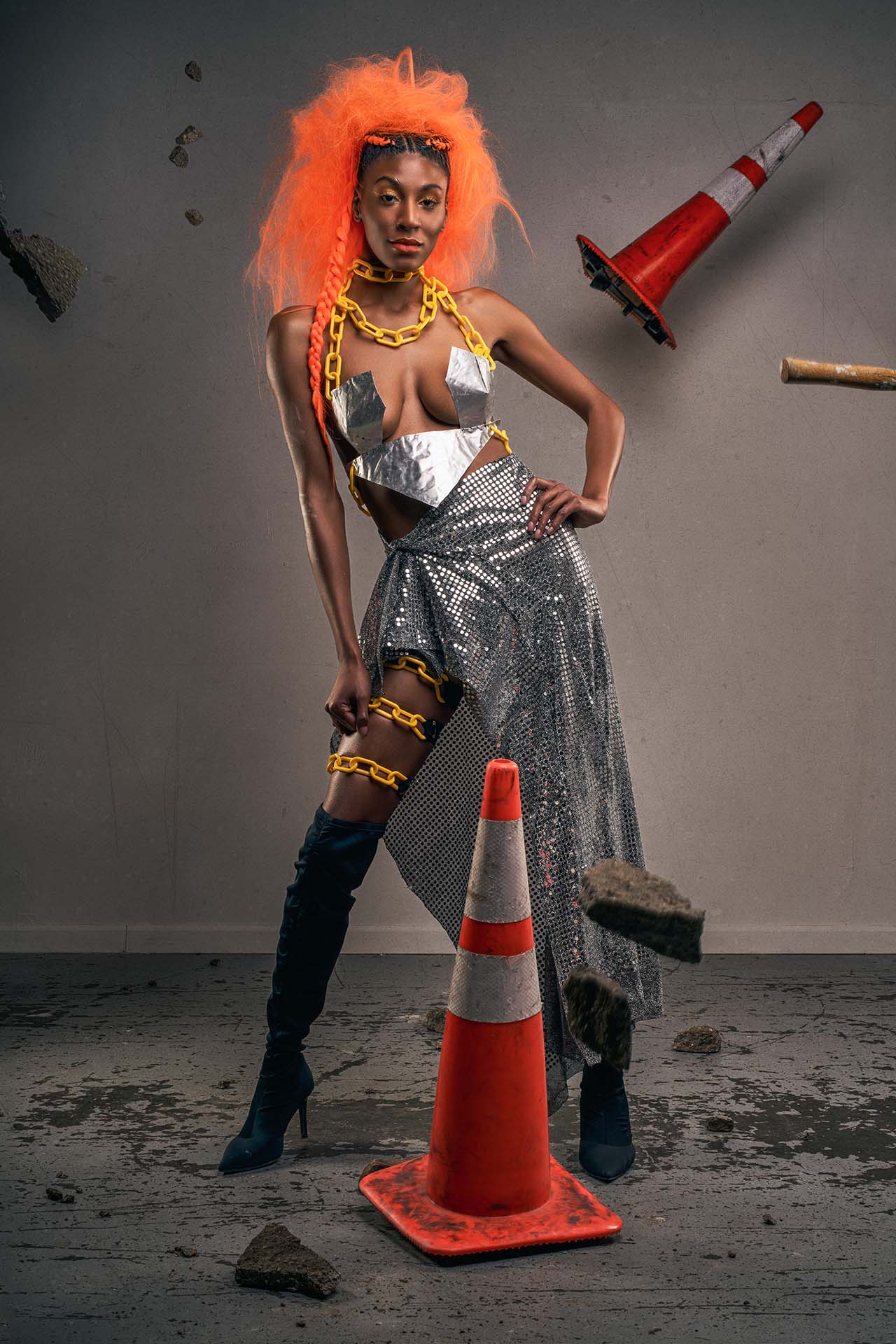 Model with an orange wig and yellow chain jewelry, wearing a metallic silver top and skirt, standing among traffic cones and rubble