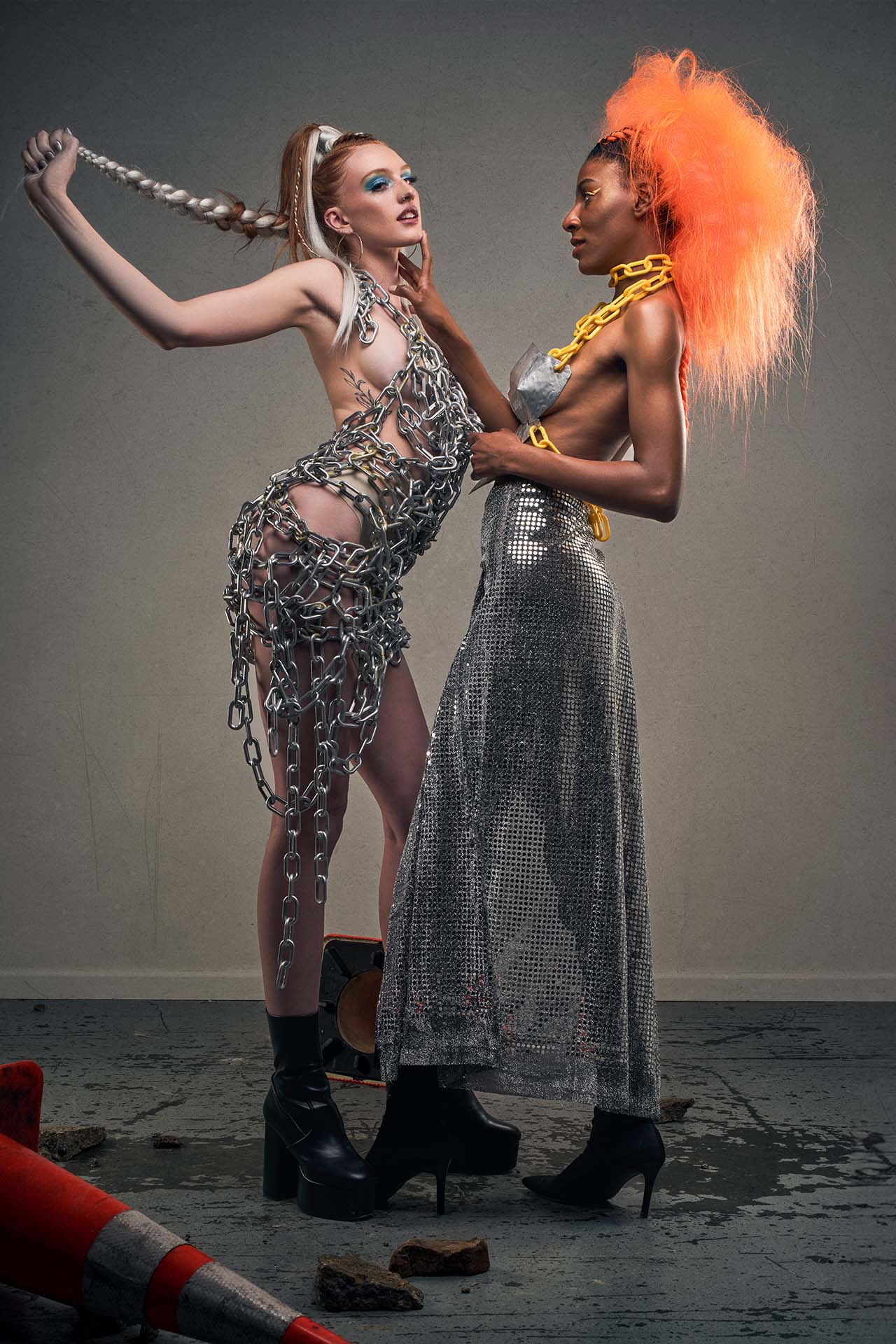 Two models in chain-link and metallic silver outfits, one with a vivid orange wig and gold chain jewelry, avant-garde editorial photography