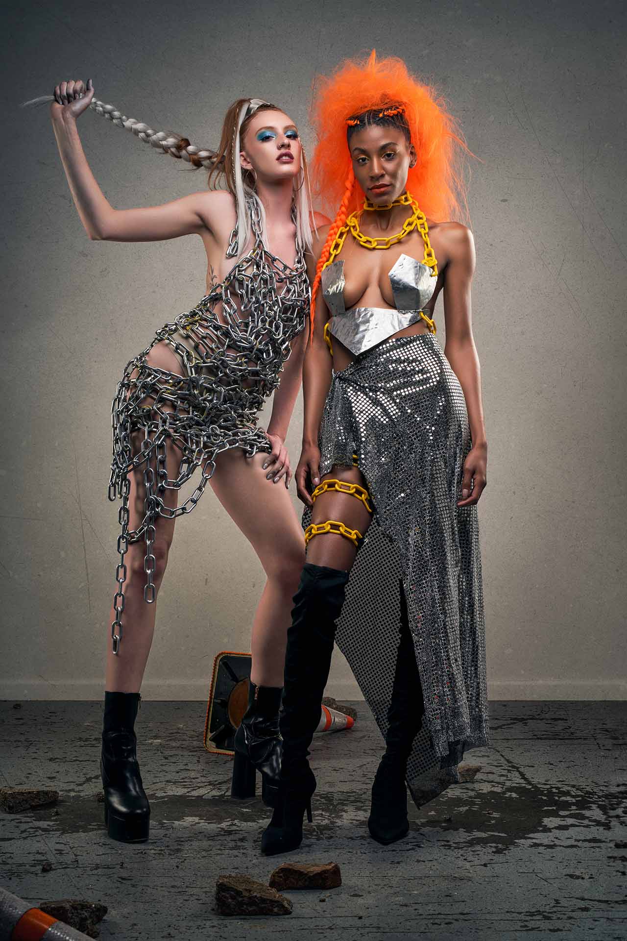 Two models in chain-link and metallic outfits posing beside a traffic cone, one with a vivid orange wig, avant-garde editorial photography