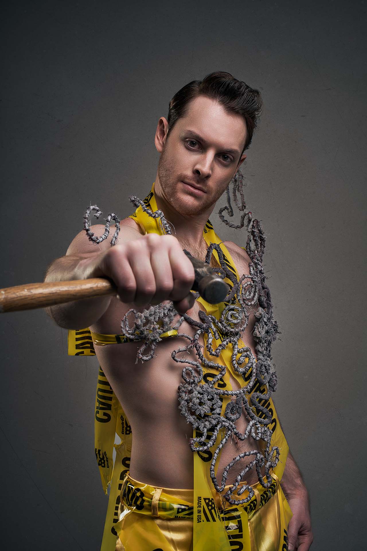 Male model in caution-tape wrapped garments holding a mallet, avant-garde construction-themed editorial
