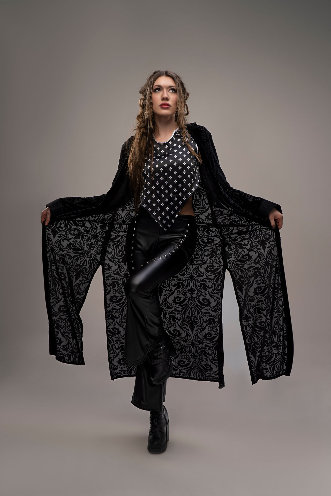 Model in a black velvet patterned coat worn open, arms spread, over a black star-print top and leather pants
