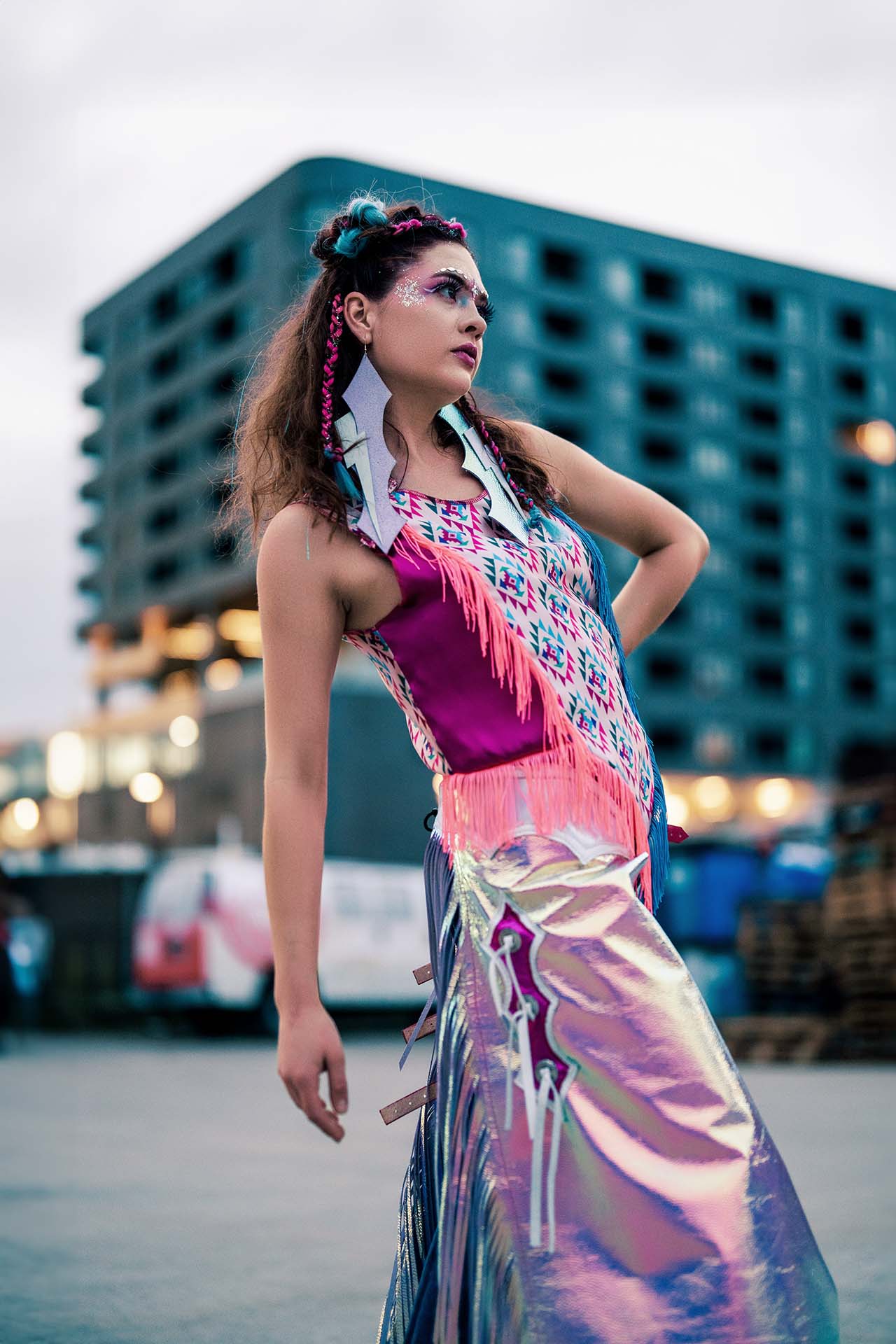 Model in a colorful fringe outfit standing on an urban street at night, city lights backdrop