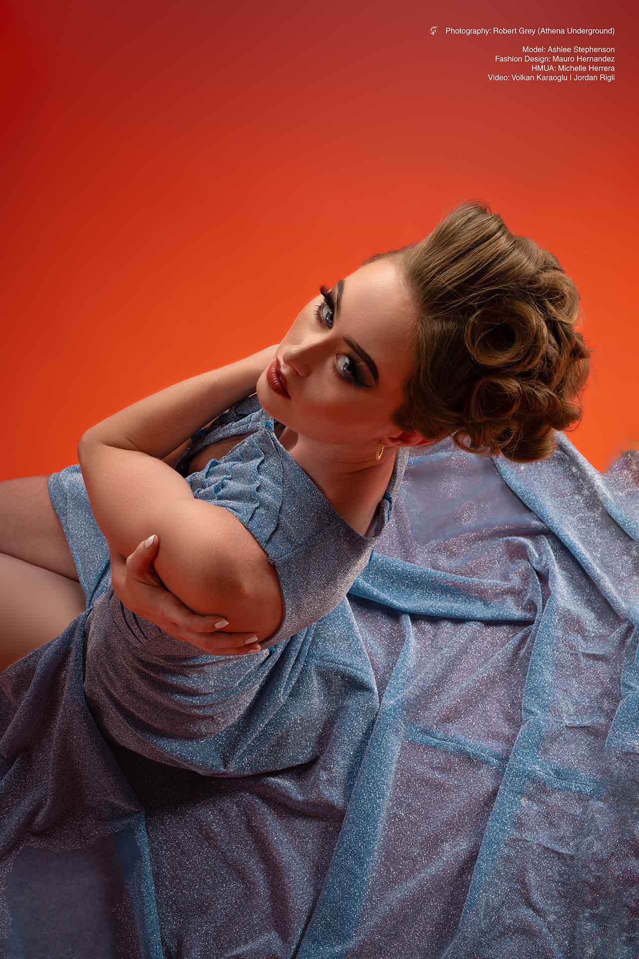 Model in a blue velour outfit seated dramatically against a red studio backdrop