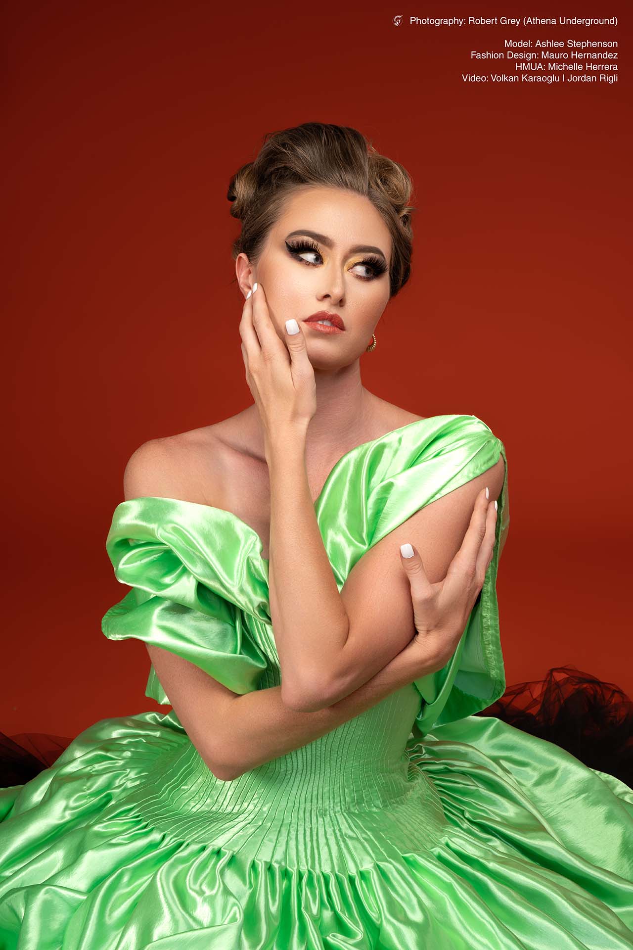 Model in a green satin off-shoulder gown, seated with hand near her face, red studio backdrop