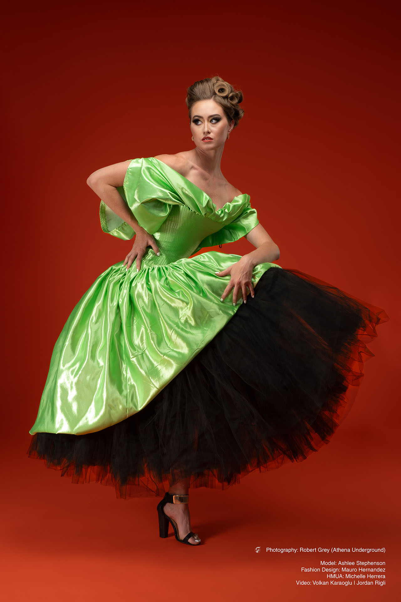 Model in a green satin gown with a black tulle underskirt, standing against a red backdrop