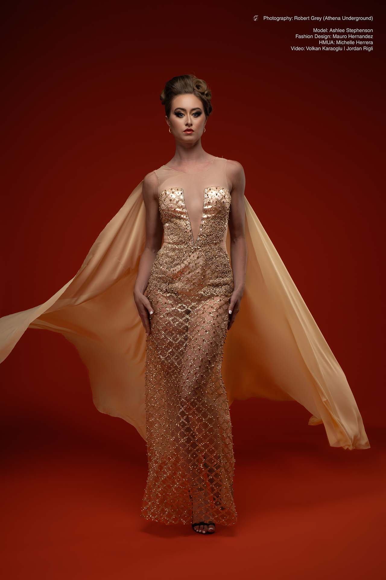 Model in a gold corset gown with a flowing cape against a red studio backdrop