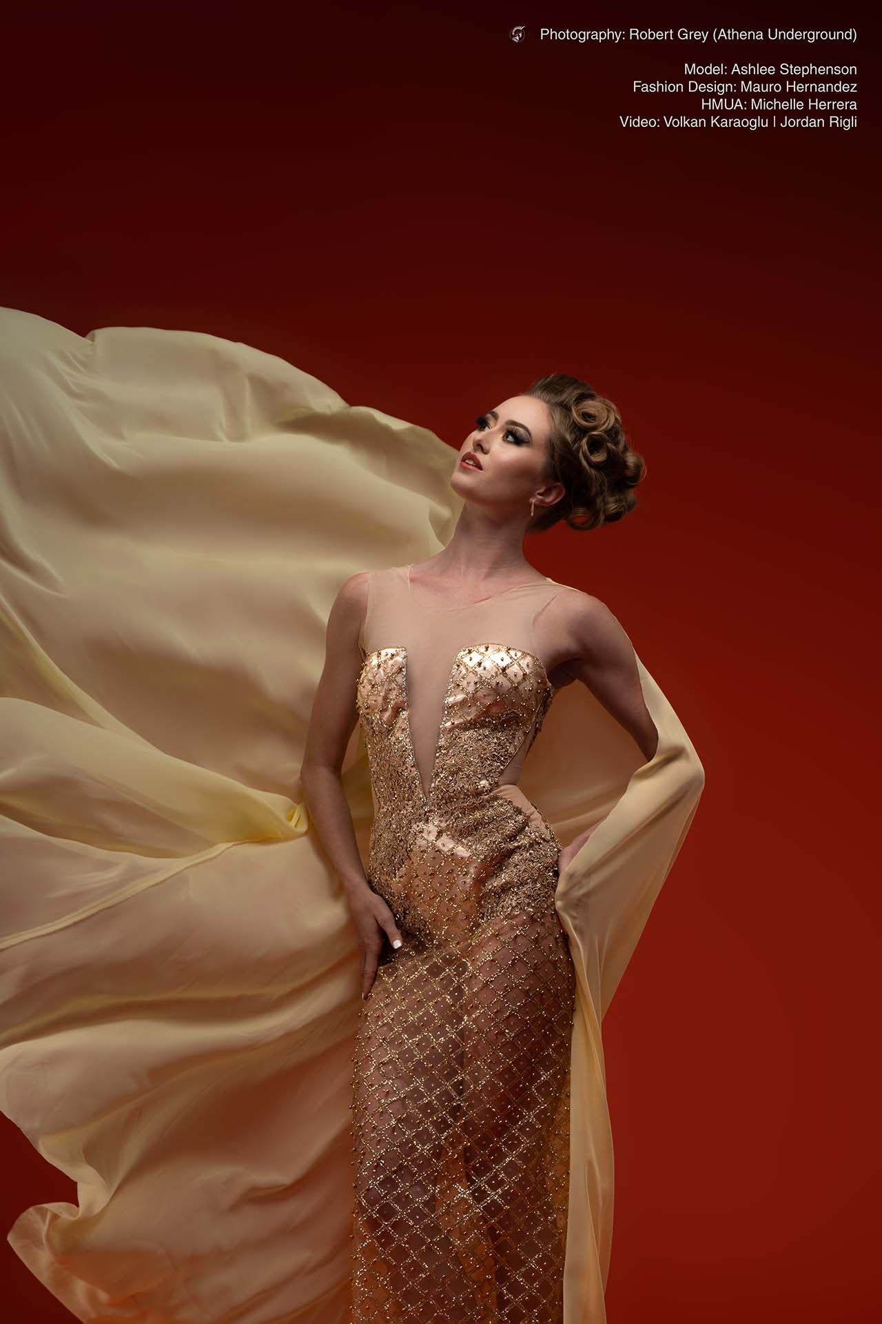 Model in a gold corset gown with flowing fabric, standing pose against a red backdrop