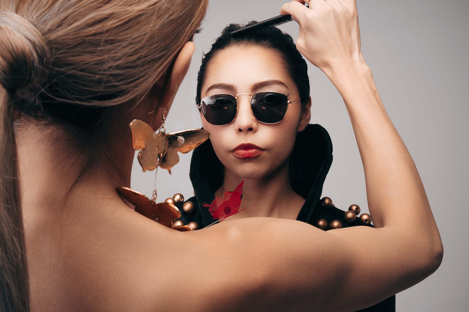 Mauro Look Book Behind-the-Scenes Photography 01 Stylist adjusting a model's hair with a comb while she poses in sunglasses and statement jewelry, behind-the-scenes styling photo