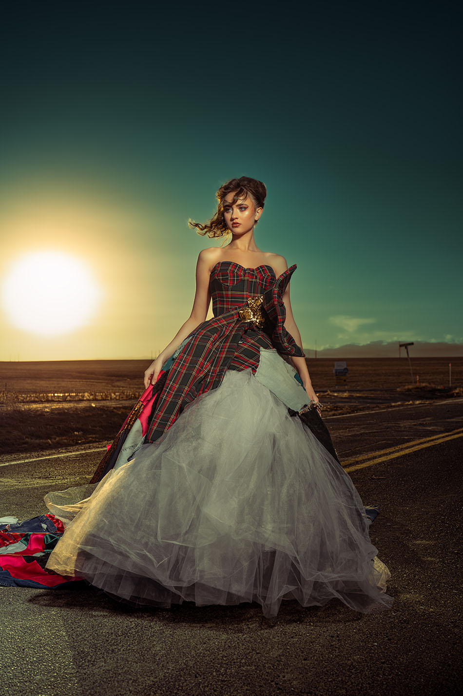 Model in a plaid corset gown with a white tulle skirt standing on a deserted road at sunset