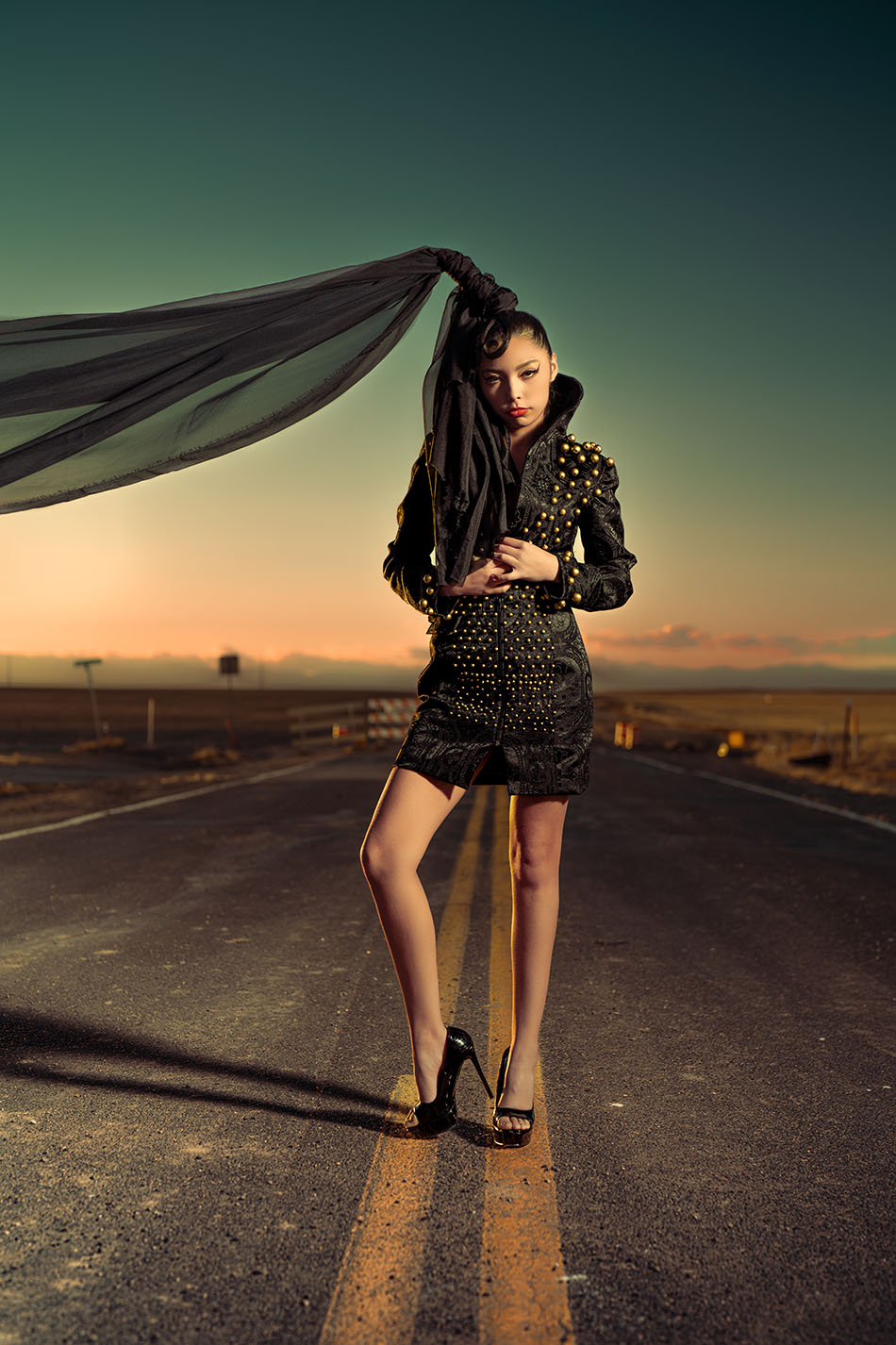 Studded Veil Editorial Photography 02 Model in a black studded outfit with a flowing veil, standing on a deserted road at sunset