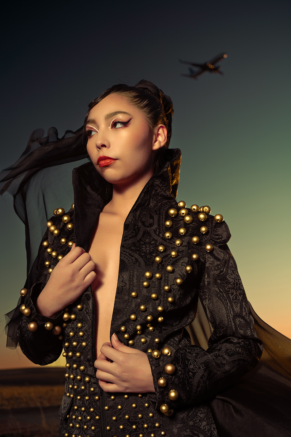 Studded Veil Editorial Photography 05 Close portrait of a model in an open gold-studded jacket with hair blowing, plane visible in the sky