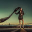 Full-length photo of a model in a black studded outfit with a flowing veil standing on a desert road