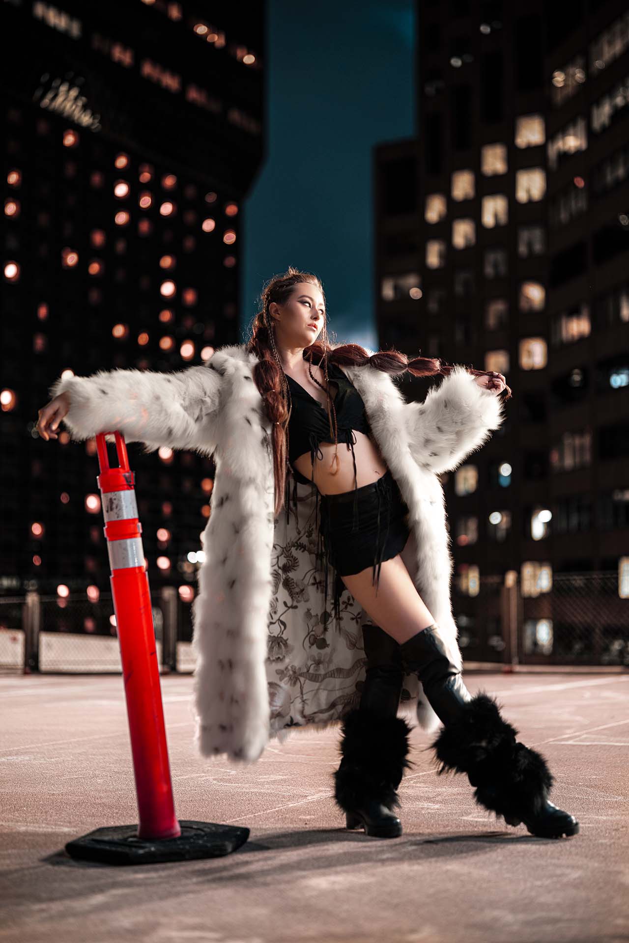 Model in fur-detailed outfit on an urban rooftop with a traffic cone prop, city skyline at dusk