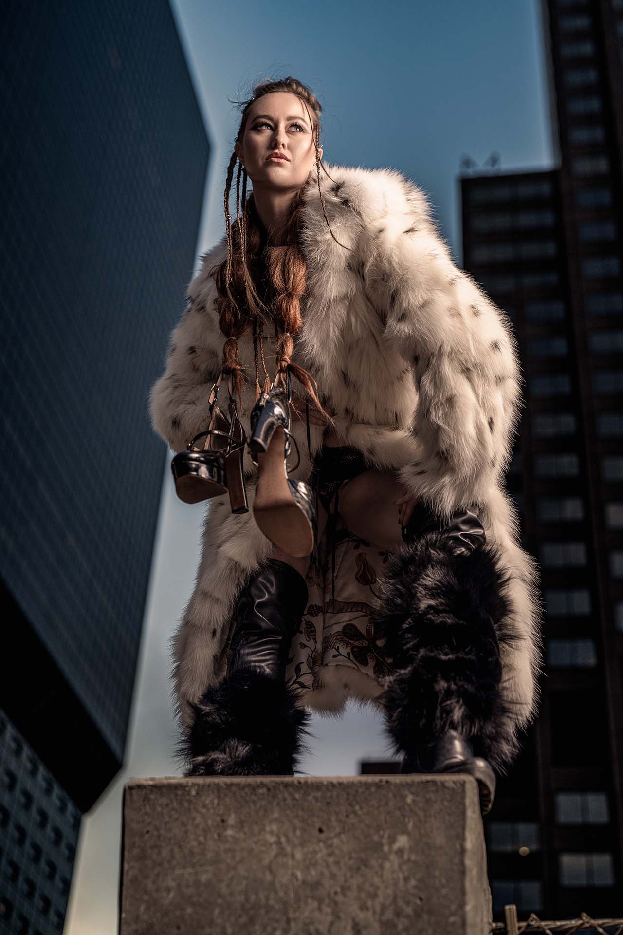 Model standing on an urban structure in a fur coat with a city backdrop, editorial fashion photography