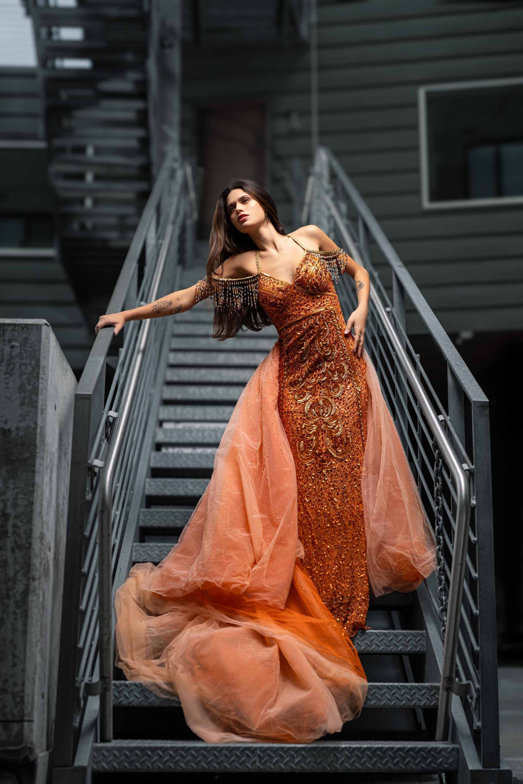 Model in a flowing orange gown captured mid-movement on a metal staircase, dramatic editorial photography