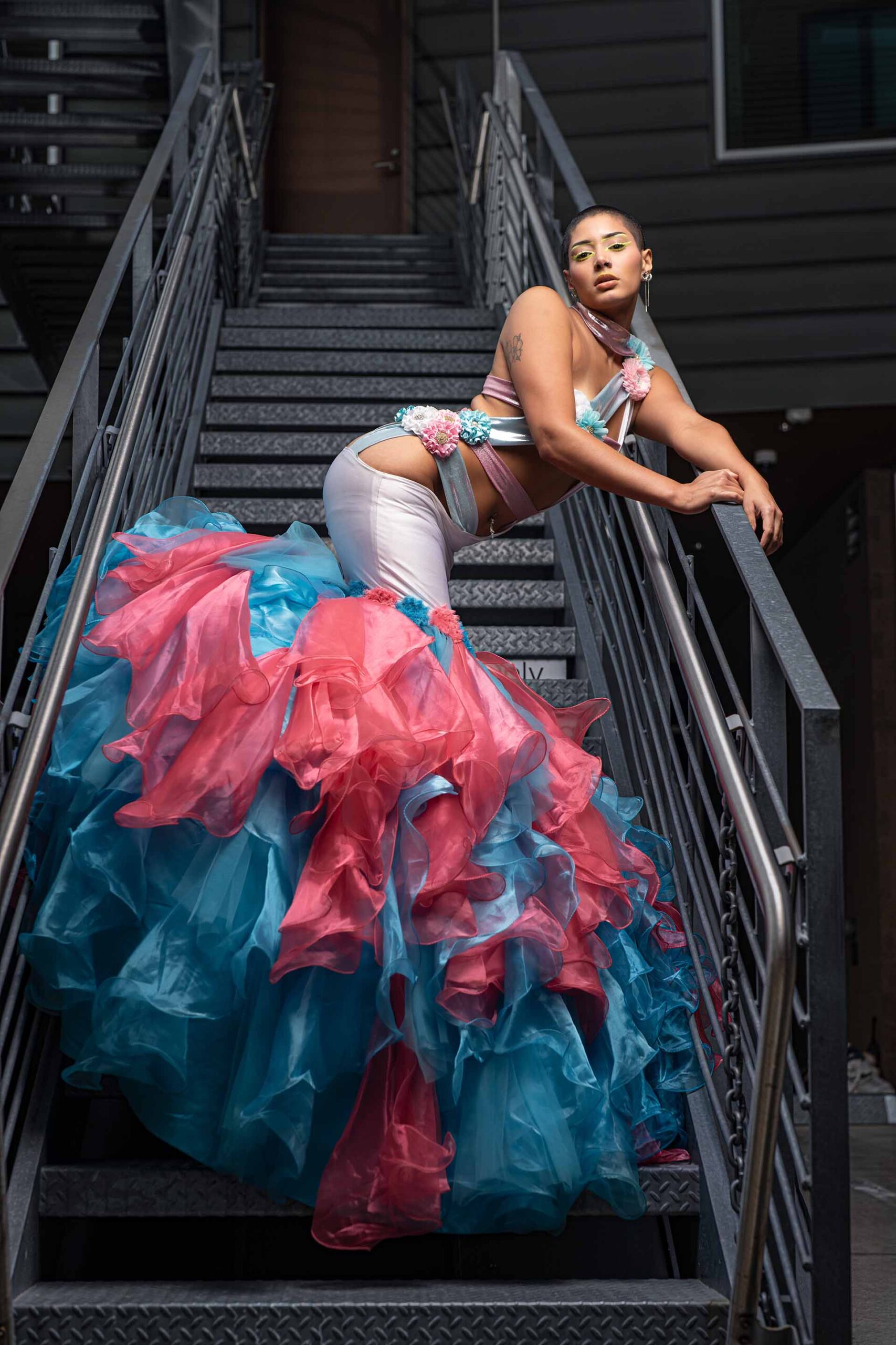 Model in a pink and teal ruffled gown posing on a metal staircase, close editorial pose