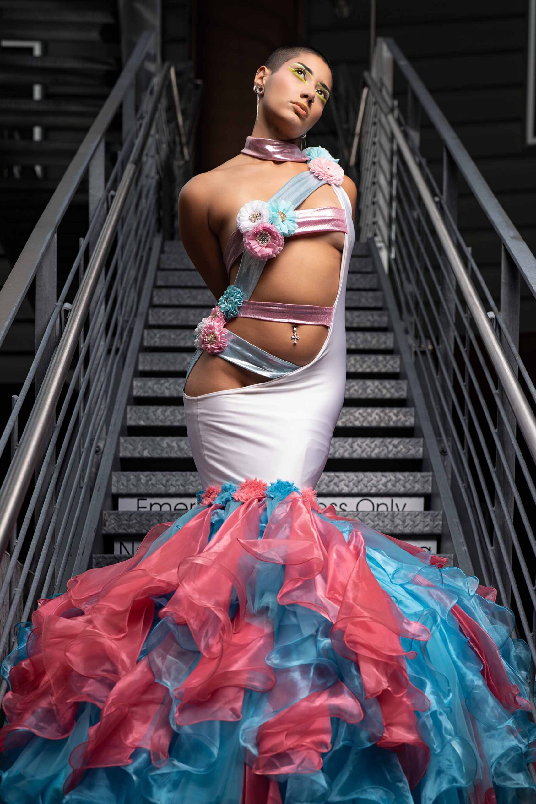 Model in a pink and teal ruffled gown on a staircase, facing forward with head tilted up and hands clasped behind her back