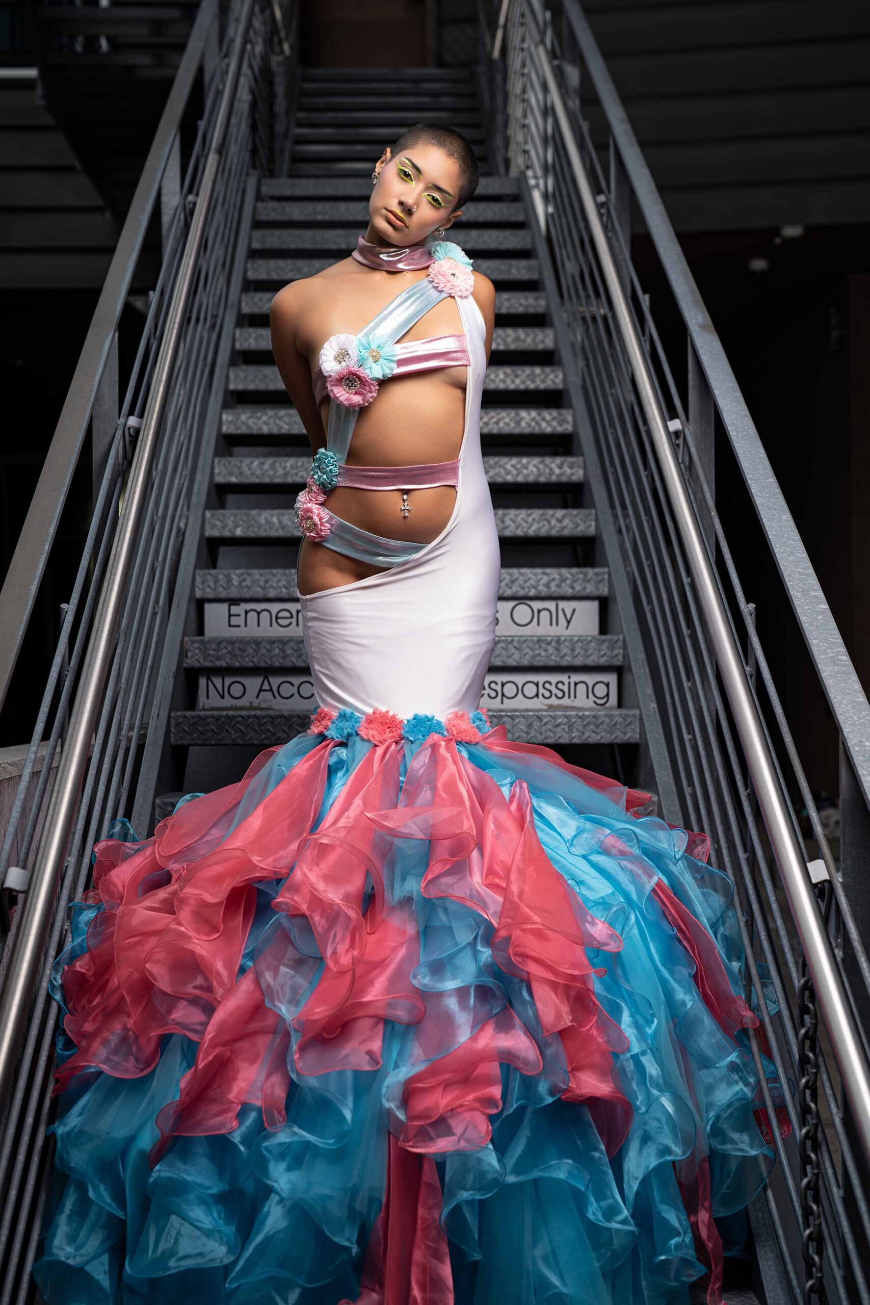 Model in a pink and teal ruffled gown on a staircase, front-facing editorial pose