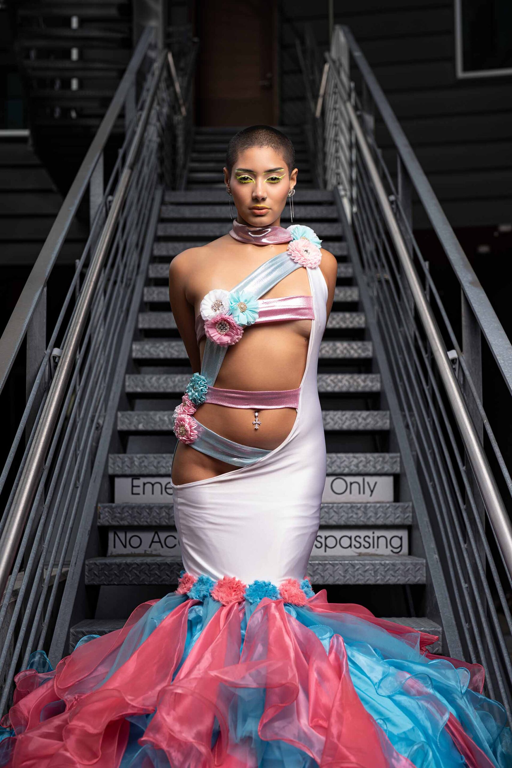 Model in a pink and teal ruffled gown standing on a metal staircase, editorial photo
