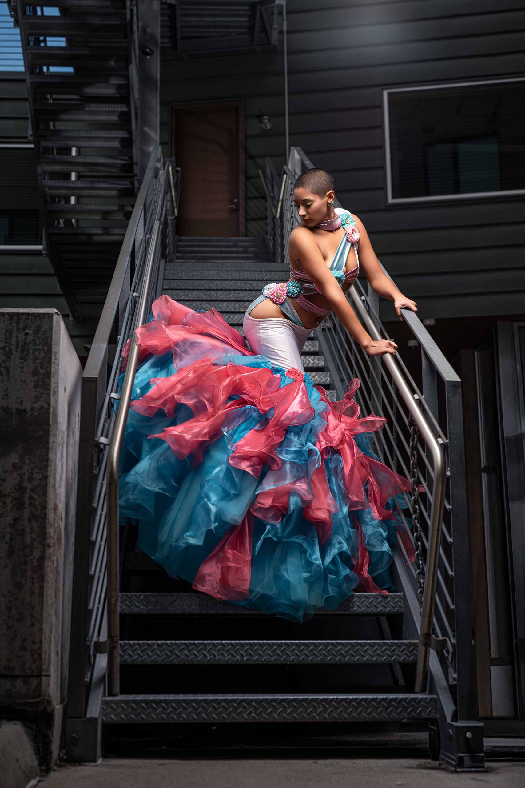 Model in a pink and teal ruffled gown on a metal staircase, editorial fashion photography