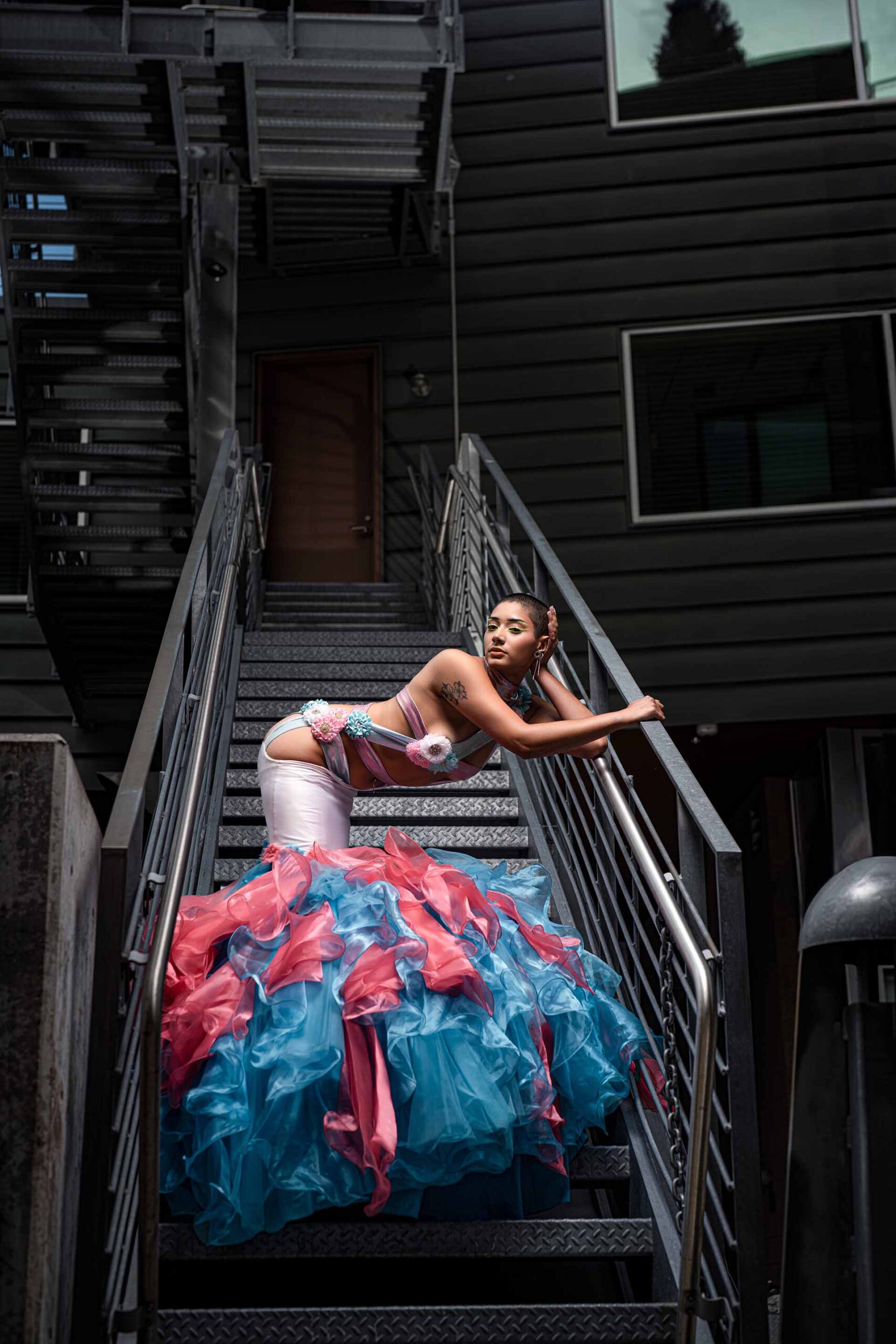 Model in a pink and teal ruffled gown leaning against a metal staircase railing outside a building, wide editorial shot
