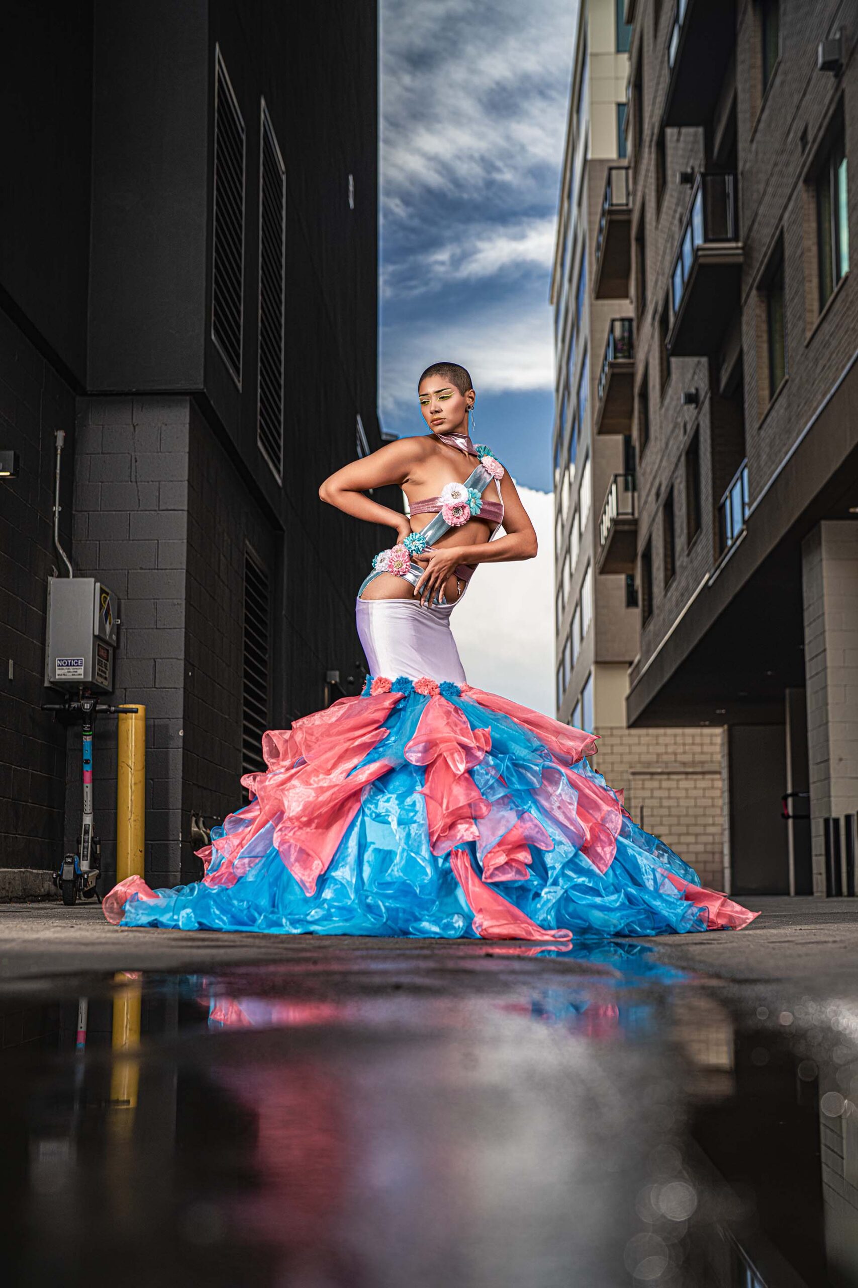 Model in a pink and teal ruffled gown standing on a wet street with reflective puddles, urban editorial photography