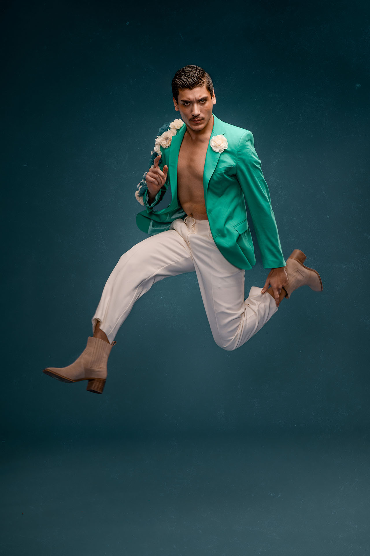 Male model in a green blazer and white pants captured mid-jump against a teal studio backdrop