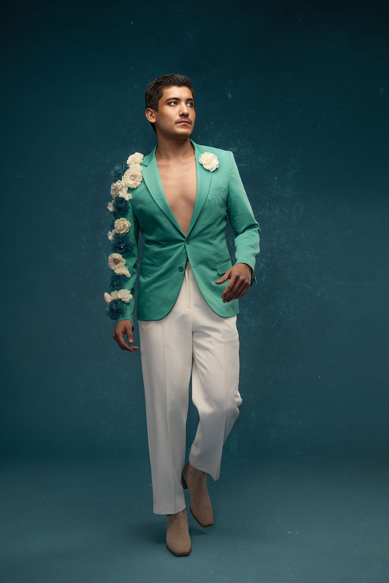 Male model in a green blazer and white pants walking against a teal studio backdrop