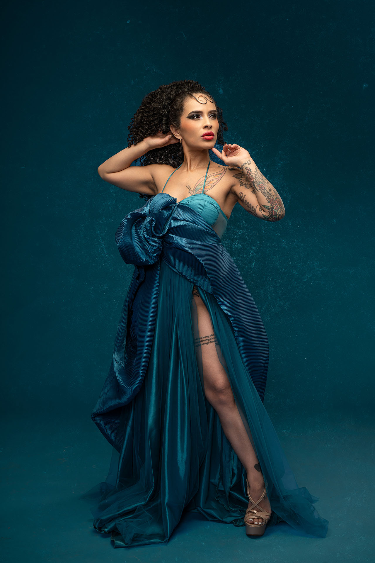 Model in a teal high-slit gown posing dramatically against a matching teal studio backdrop