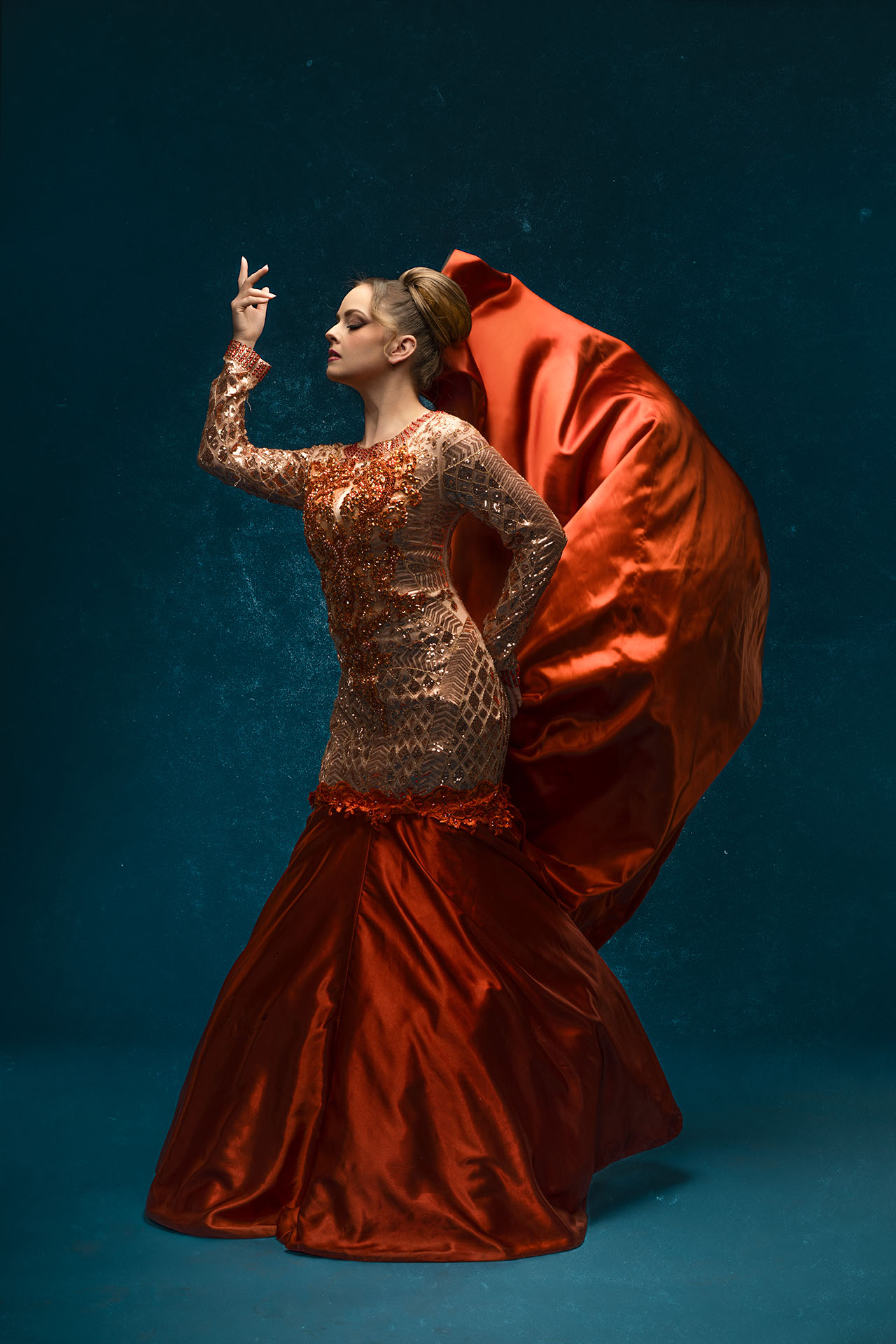 Model in a gold beaded gown with the orange skirt thrown into motion, teal studio backdrop