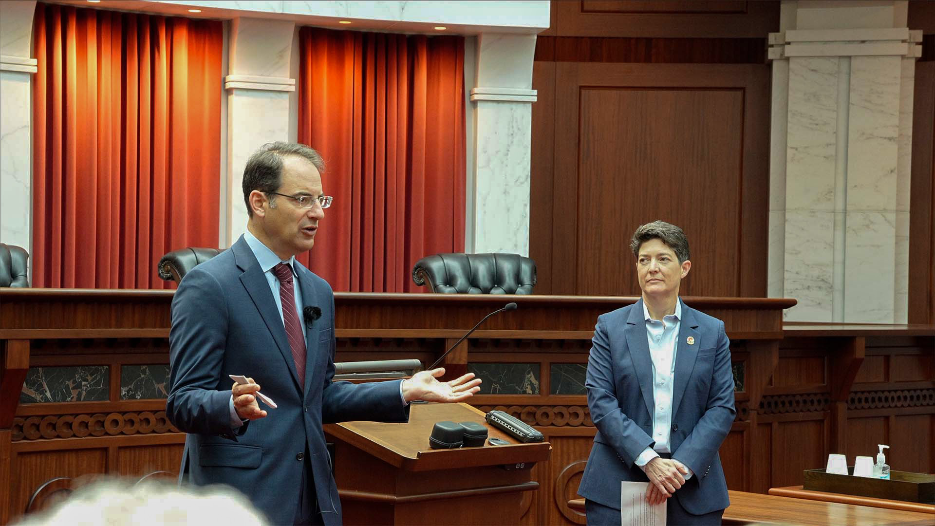 Colorado Attorney General Phil Weiser speaking beside Chief Justice Monica Márquez in a wood-paneled courtroom at an ABOTA event