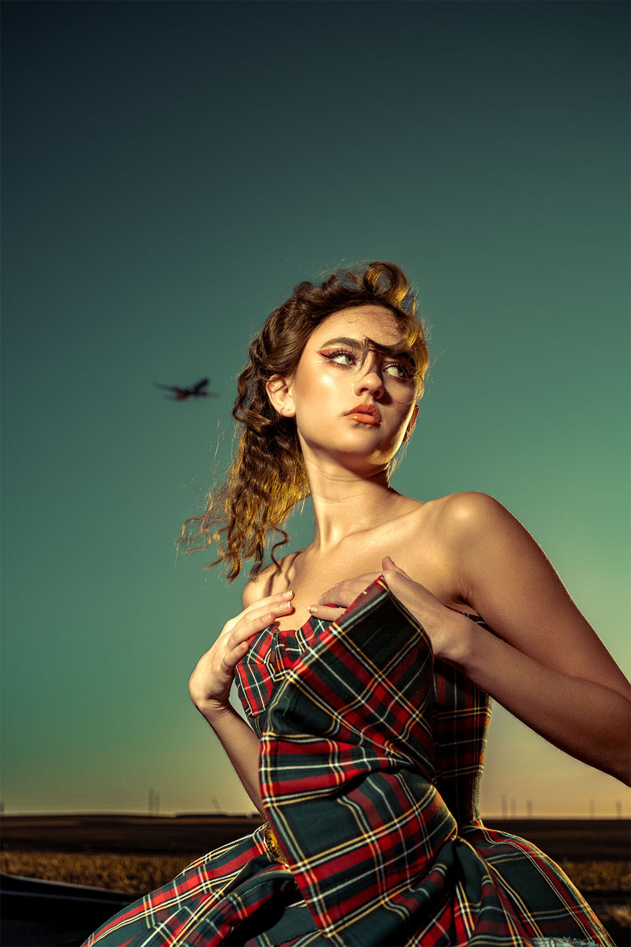 Model in a plaid corset top posing against a dramatic sky with a plane overhead, editorial fashion photography