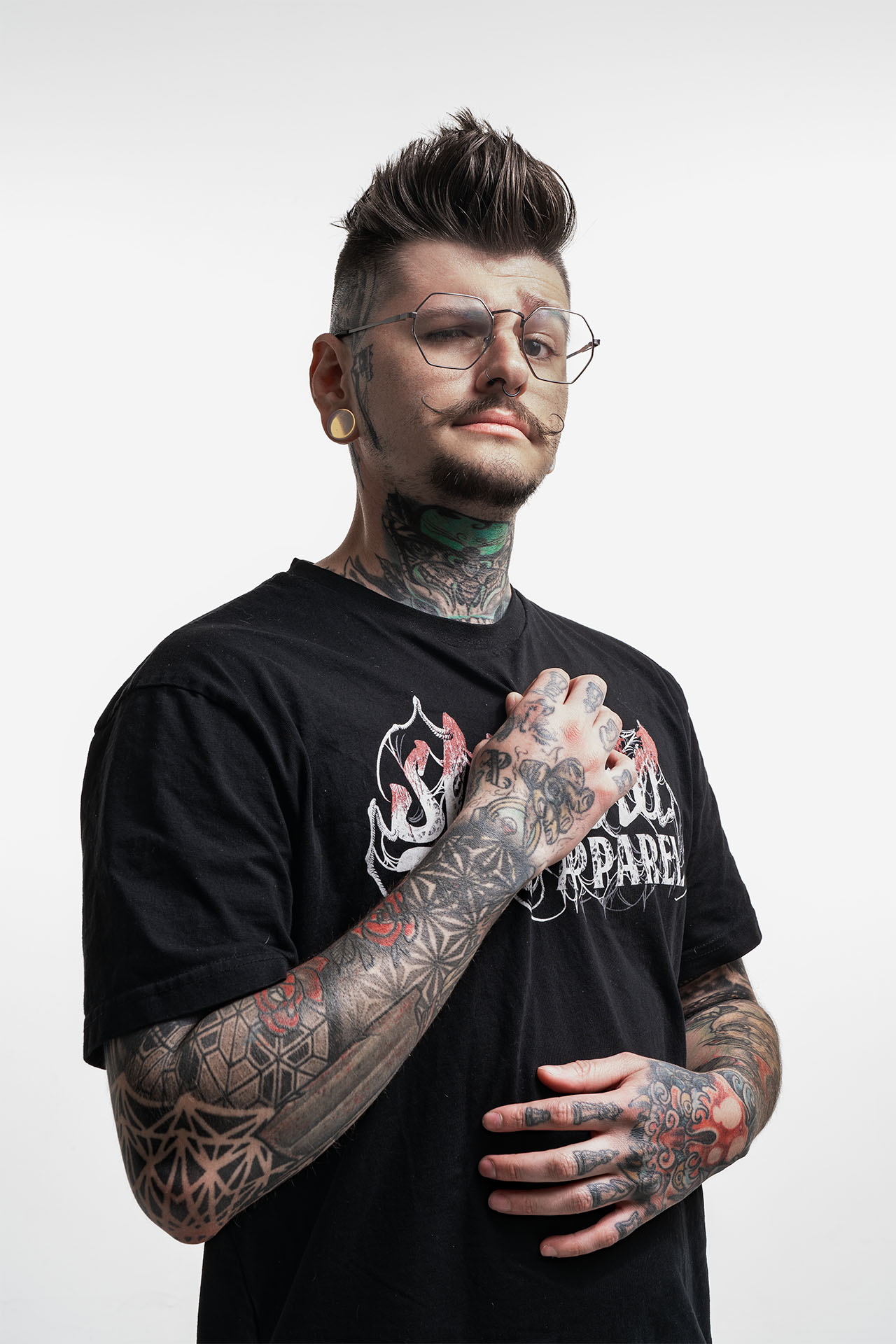 Portrait of a tattoo artist with visible tattoos, glasses, and a tattoo shop branded shirt