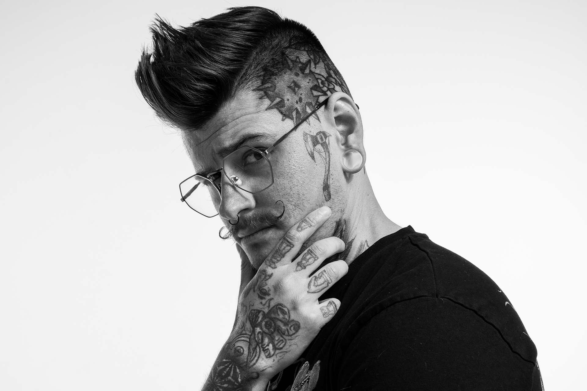 Black-and-white profile portrait of a man with tattoos, hand resting near his face